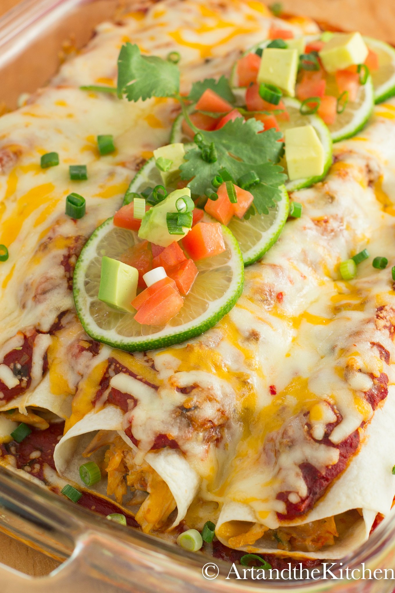 Baking dish of chicken Enchiladas topped with melted cheese, lime slices and chucks of avocado, tomatoes and green onions.