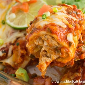 Baking dish of chicken Enchiladas topped with melted cheese, lime slices and chucks of avocado, tomatoes and green onions.