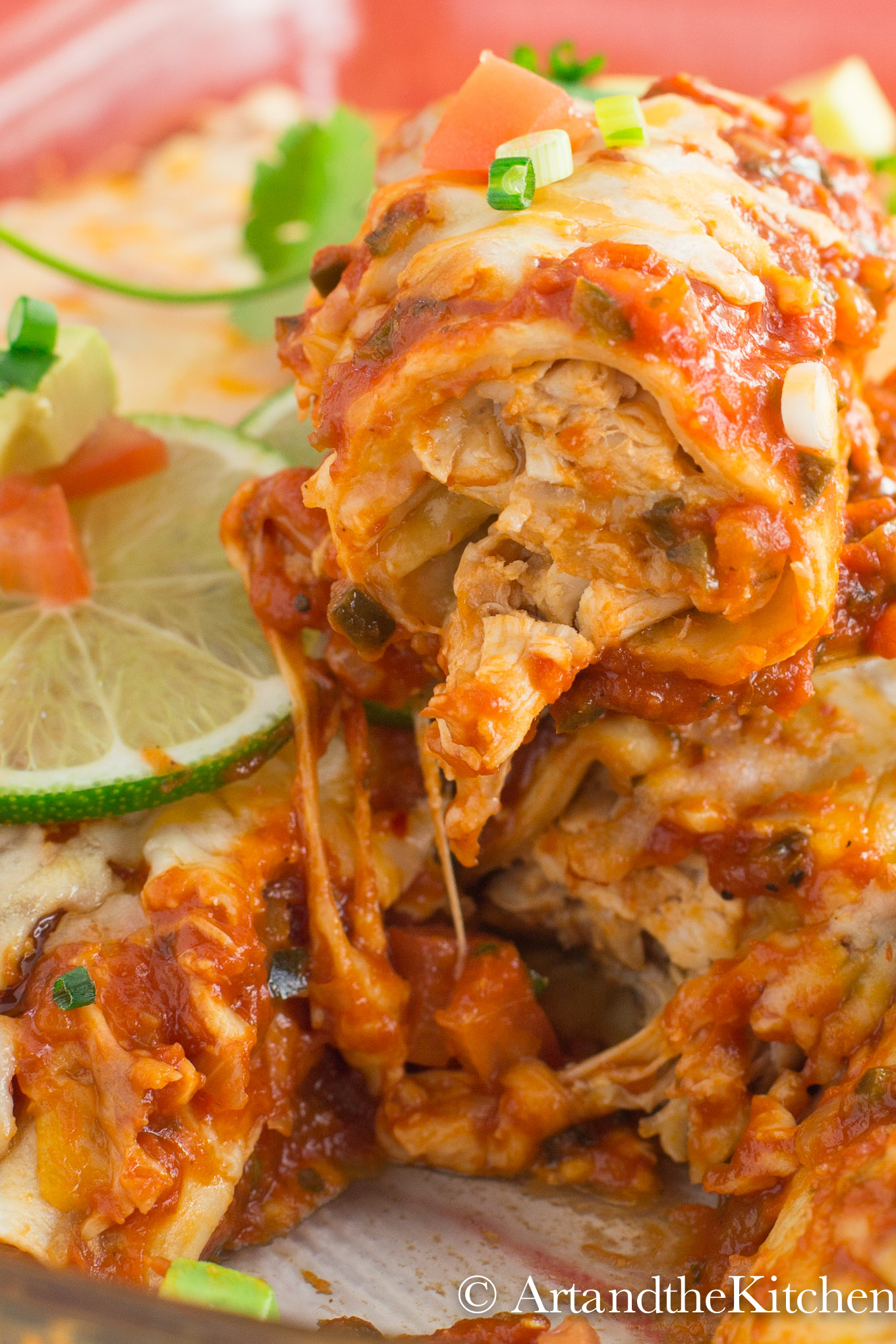 Baking dish of chicken Enchiladas topped with melted cheese, lime slices and chucks of avocado, tomatoes and green onions.