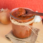 Wooden spoon scooping enchilada sauce from glass jar filled with enchilada sauce.