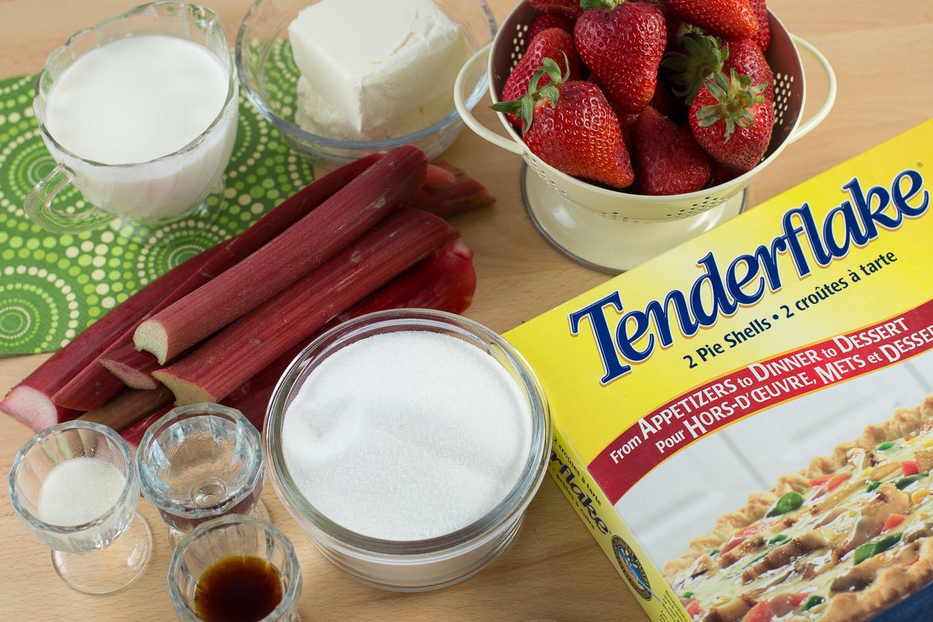 Ingredients on wood cutting board, fresh rhubarb, strawberries, pie crust, bowls of sugar, cream, butter and spices.