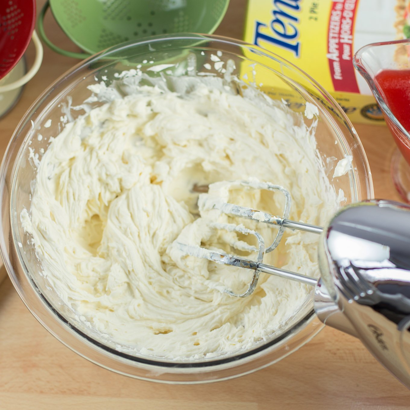 Hand mixer beating cream cheese in glass bowl.