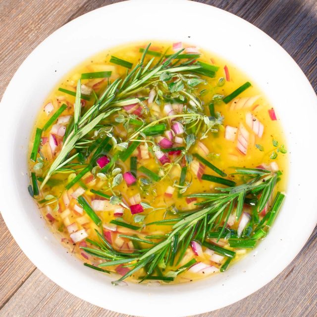 Marinade of red onions, oil and rosemary sprigs in white bowl.