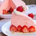 A slice of fluffy no bake strawberry pie, fresh strawberries on white plate with whole pie with slice cut out in background.