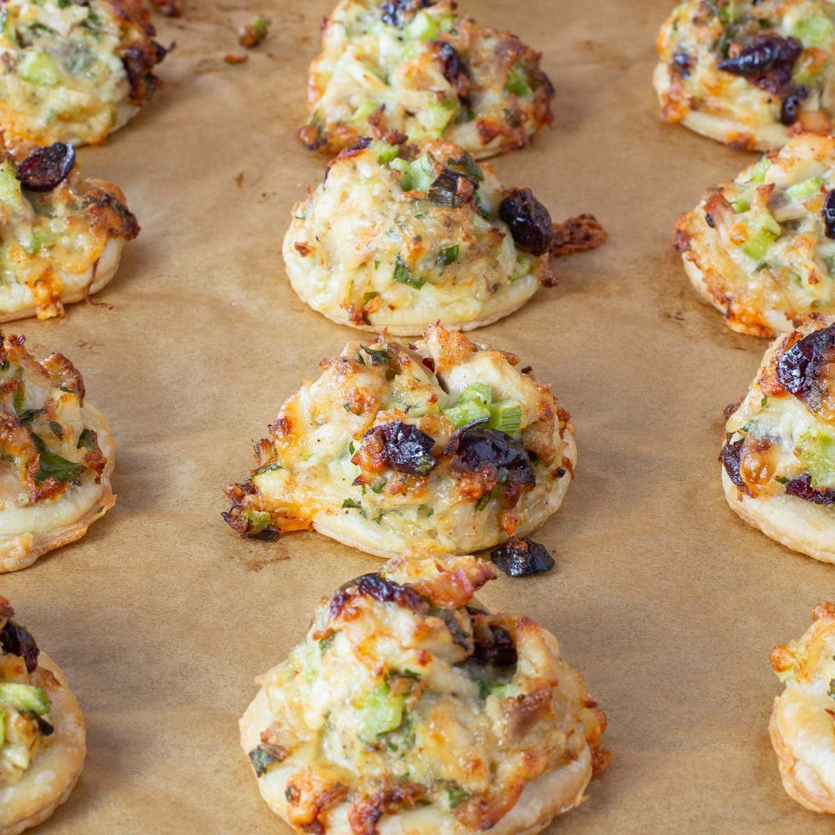 Rows of small, round pastries topped with chopped turkey, melted cheese, celery, green onions, and cranberries on brown parchment paper.