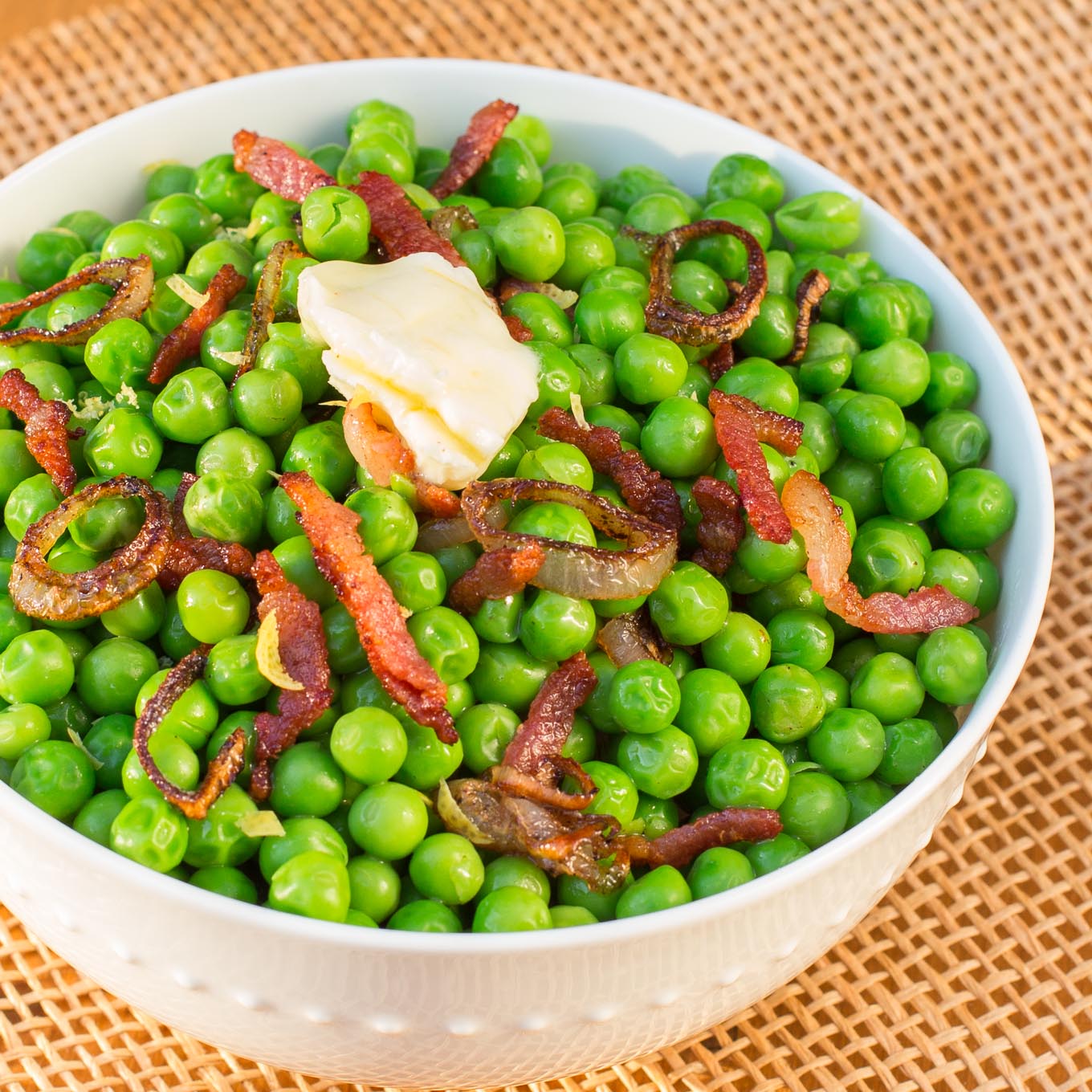 White bowl filled with green peas mixed with bacon and shallots. Topped with a pat of butter.