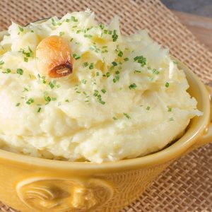 Yellow bowl filled with mashed potatoes, topped with clove of roasted garlic.