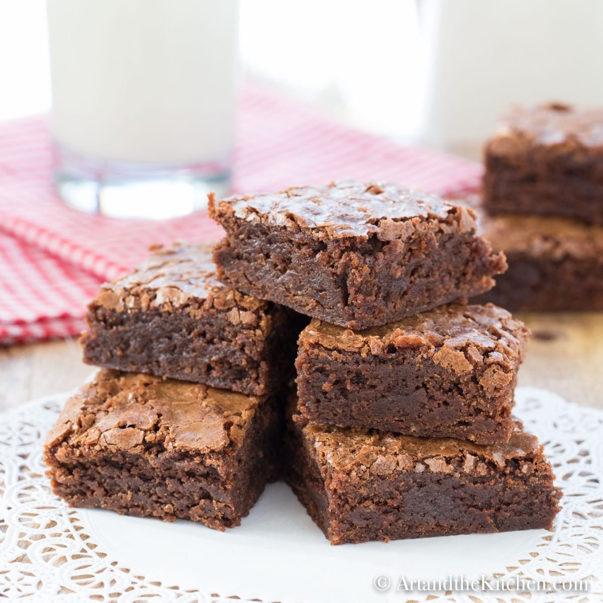A stack of five rich, fudgy brownies with a crackly top sits on a doily-covered plate. Background includes a glass of milk and a red gingham cloth.