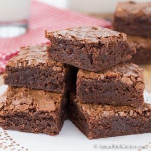 A stack of five rich, fudgy brownies with a crackly top sits on a doily-covered plate.