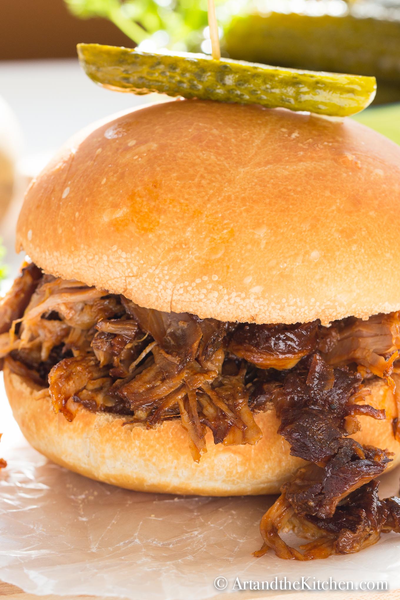 Bun filled with barbecue coated pulled pork, garnished with a dill pickle on top.