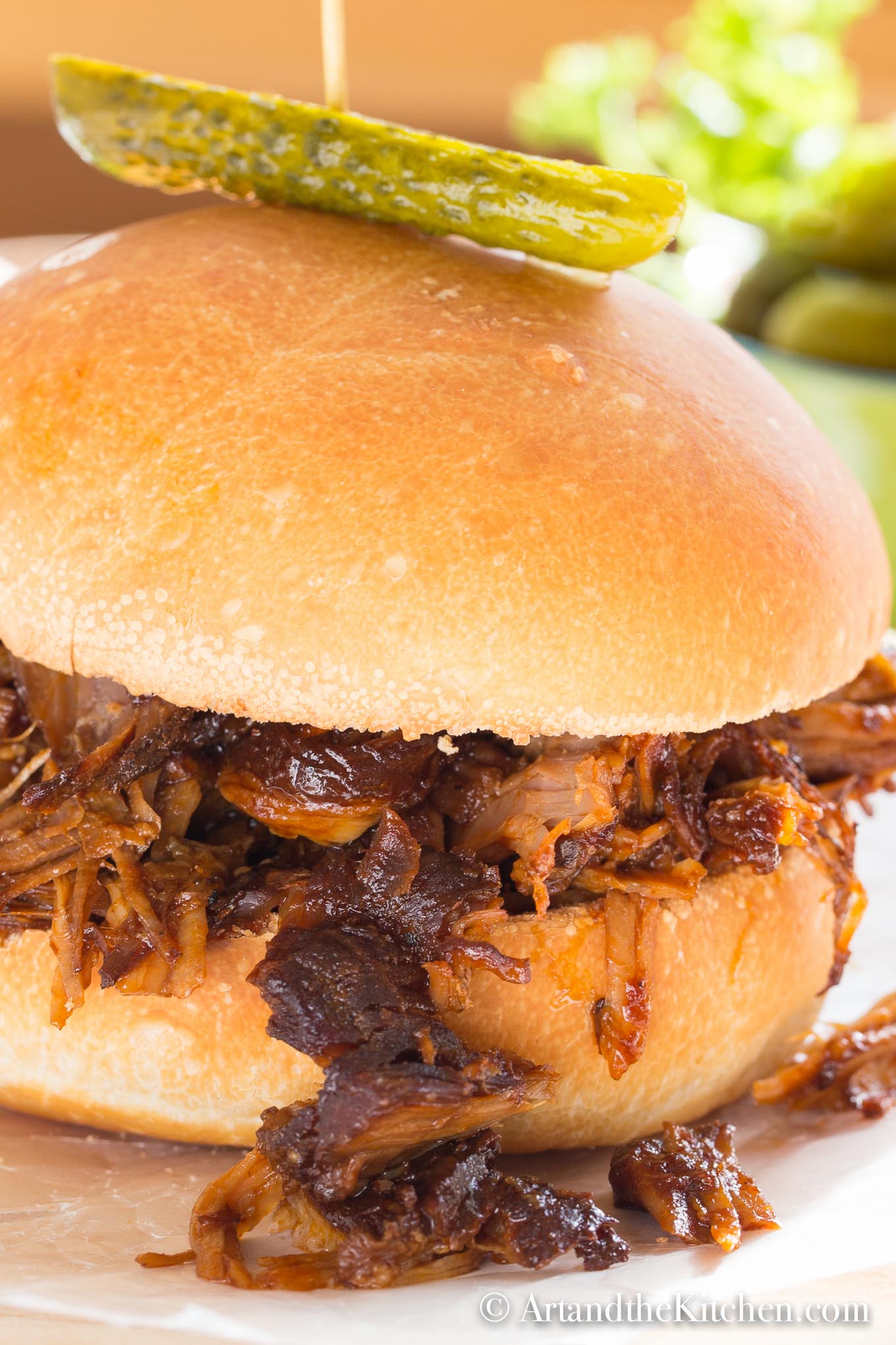 Bun filled with barbecue coated pulled pork, garnished with a dill pickle on top.