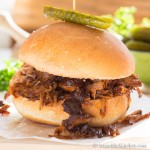 Bun filled with barbecue coated pulled pork, garnished with a dill pickle on top.