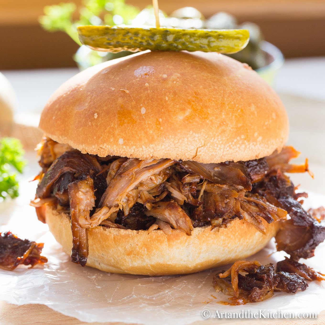 Bun filled with barbecue coated pulled pork, garnished with a dill pickle on top.