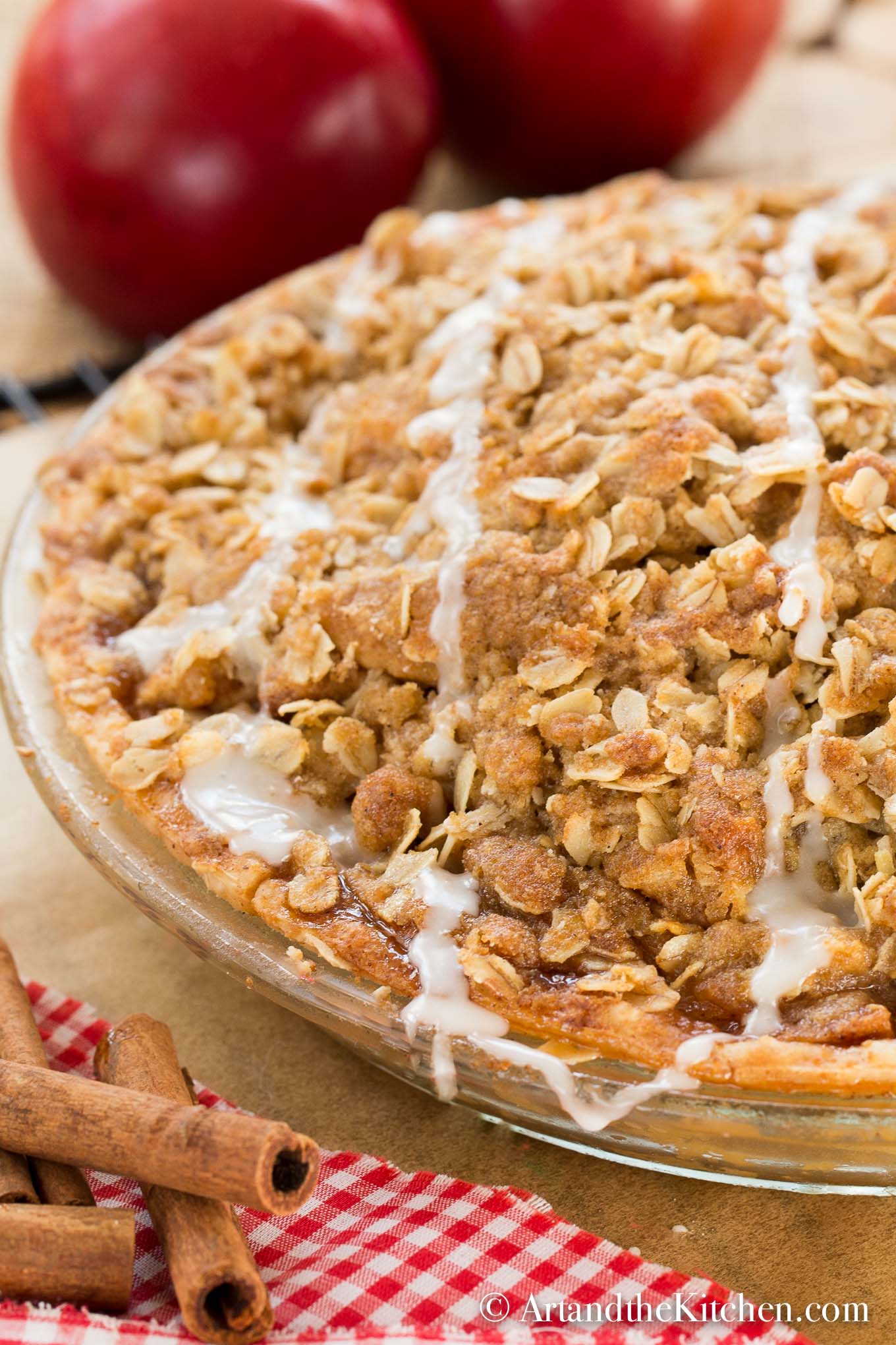 Apple pie with golden flaky crust on checkered cloth with basket of apples in background.