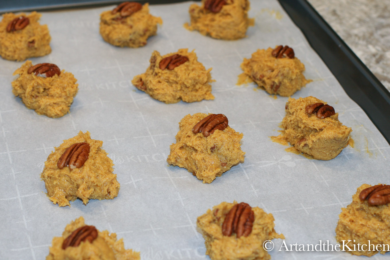 Baking sheet lined with parchment paper filled with scooped out pumpkin cookie dough. Each cookie topped with a pecan.