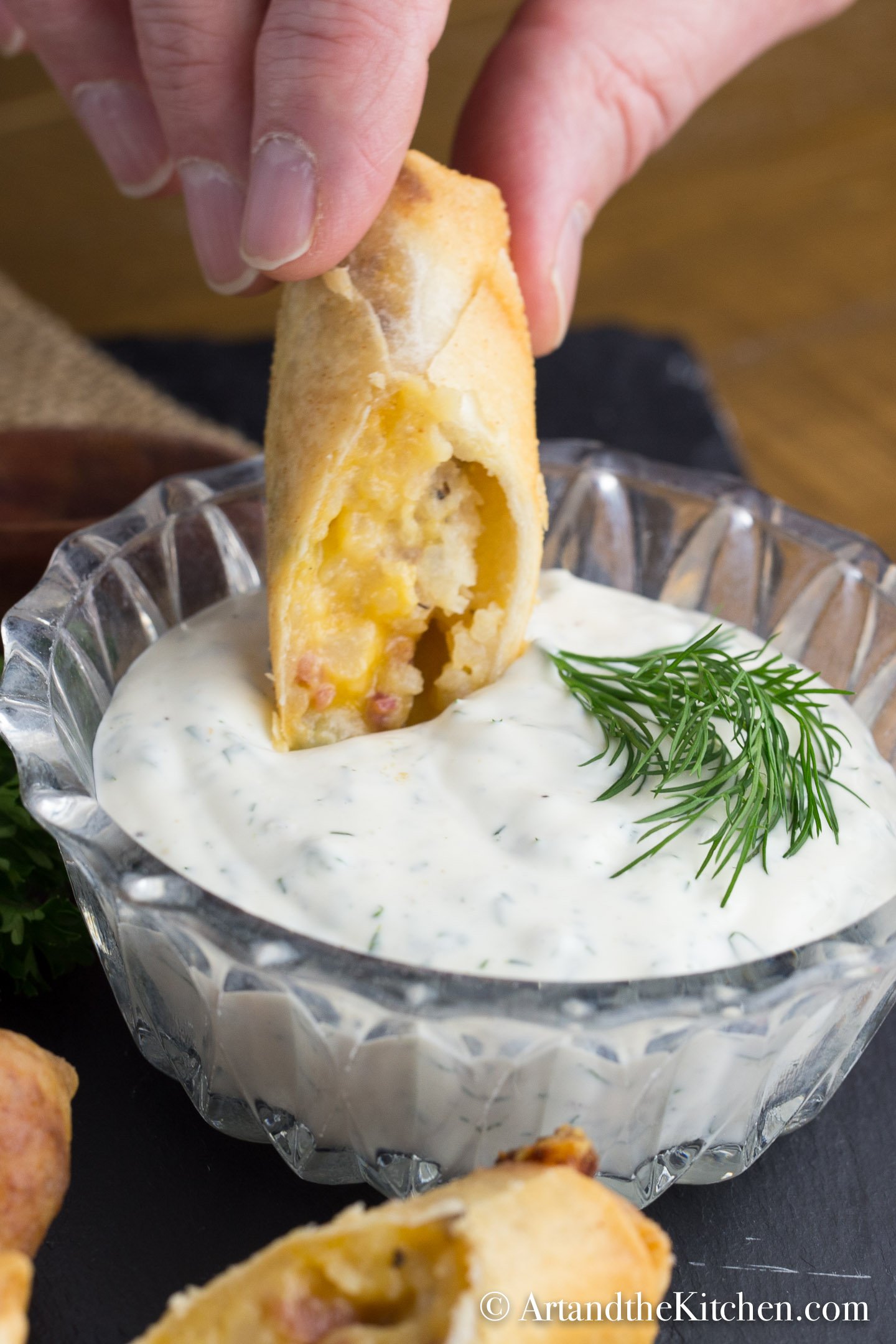 Spring roll being dipped into glass bowl filled with ranch dip. Garnished with dill sprig.