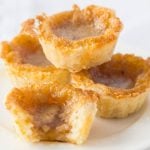 Close-up of golden-brown butter tarts with flaky crusts and gooey caramel centers, stacked on a plate.