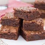 A stack of rich, fudge brownies with a crackly top sits on a paper doily-covered plate.
