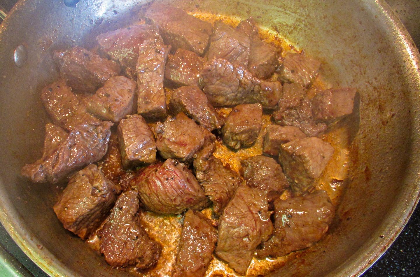 Cubes of beef searing in stainless steel frying pan.