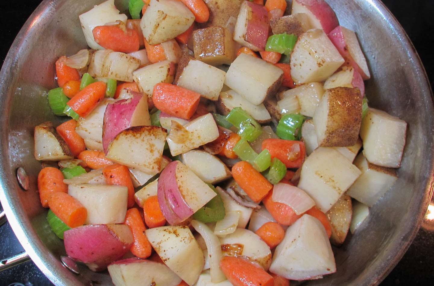 Potatoes, carrots, celery and onions searing in stainless steel skillet.