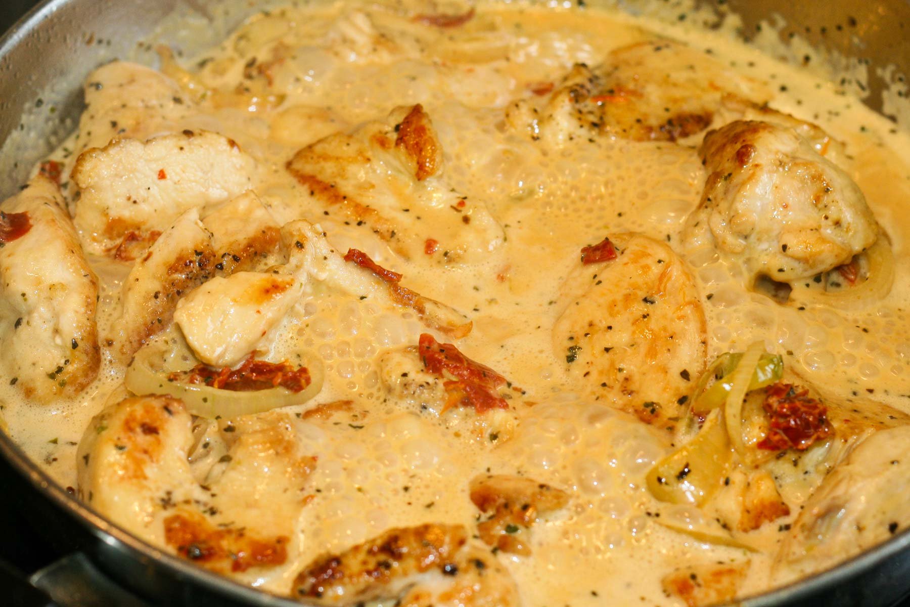 Chicken with shallots and sun-dried tomatoes in a creamy sauce simmering in a stainless steel frying pan.