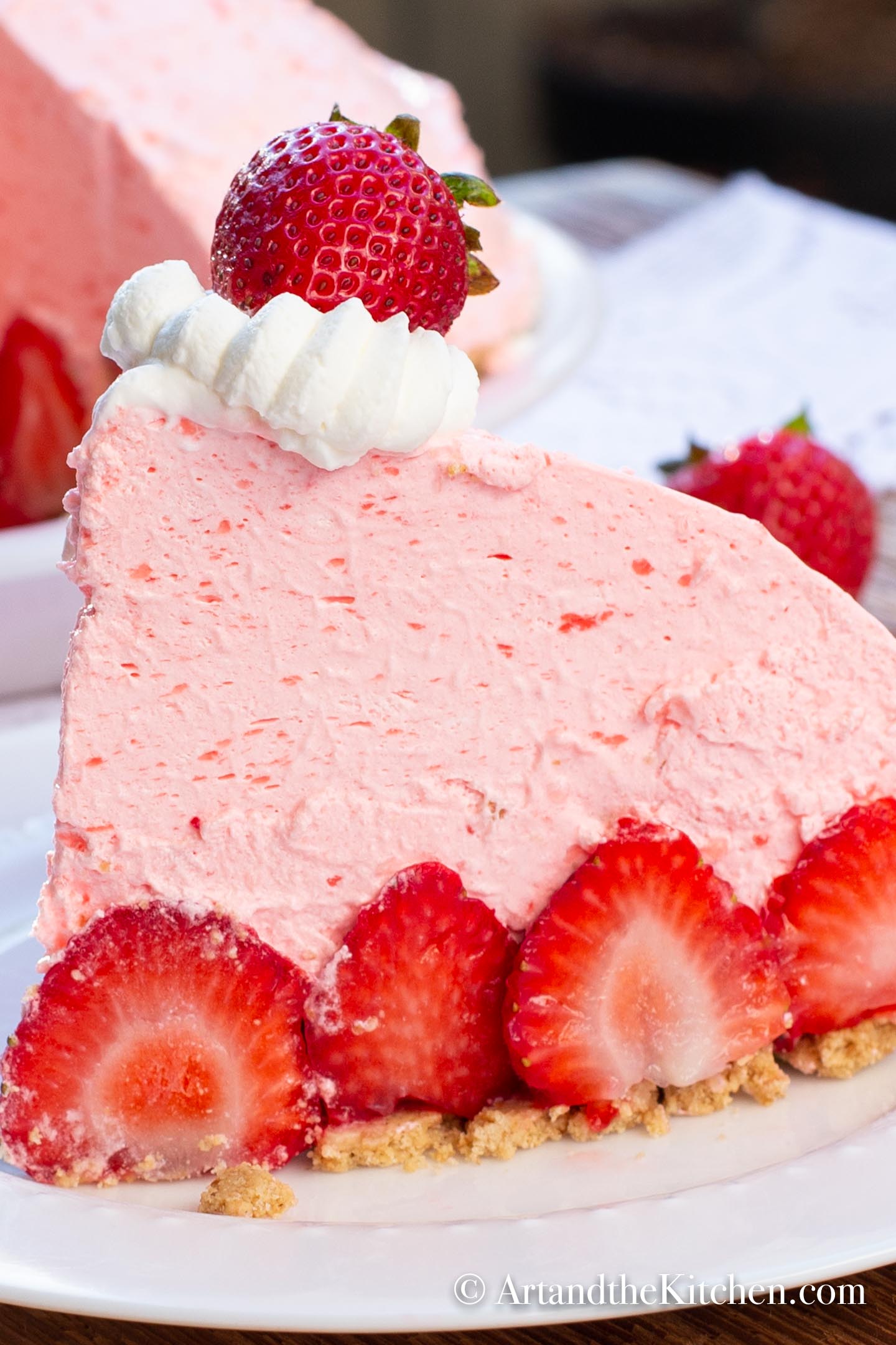 A slice of fluffy no bake strawberry pie, fresh strawberries  on white plate with whole pie with slice cut out in background.