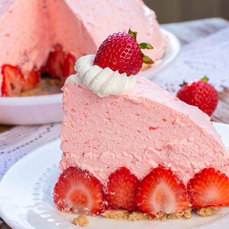 A slice of fluffy no bake strawberry pie, fresh strawberries on white plate with whole pie with slice cut out in background.