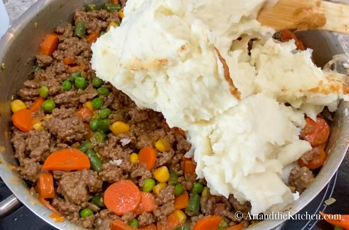 Mashed potatoes being scooped onto cooked ground beef, gravy and vegetable mixture.