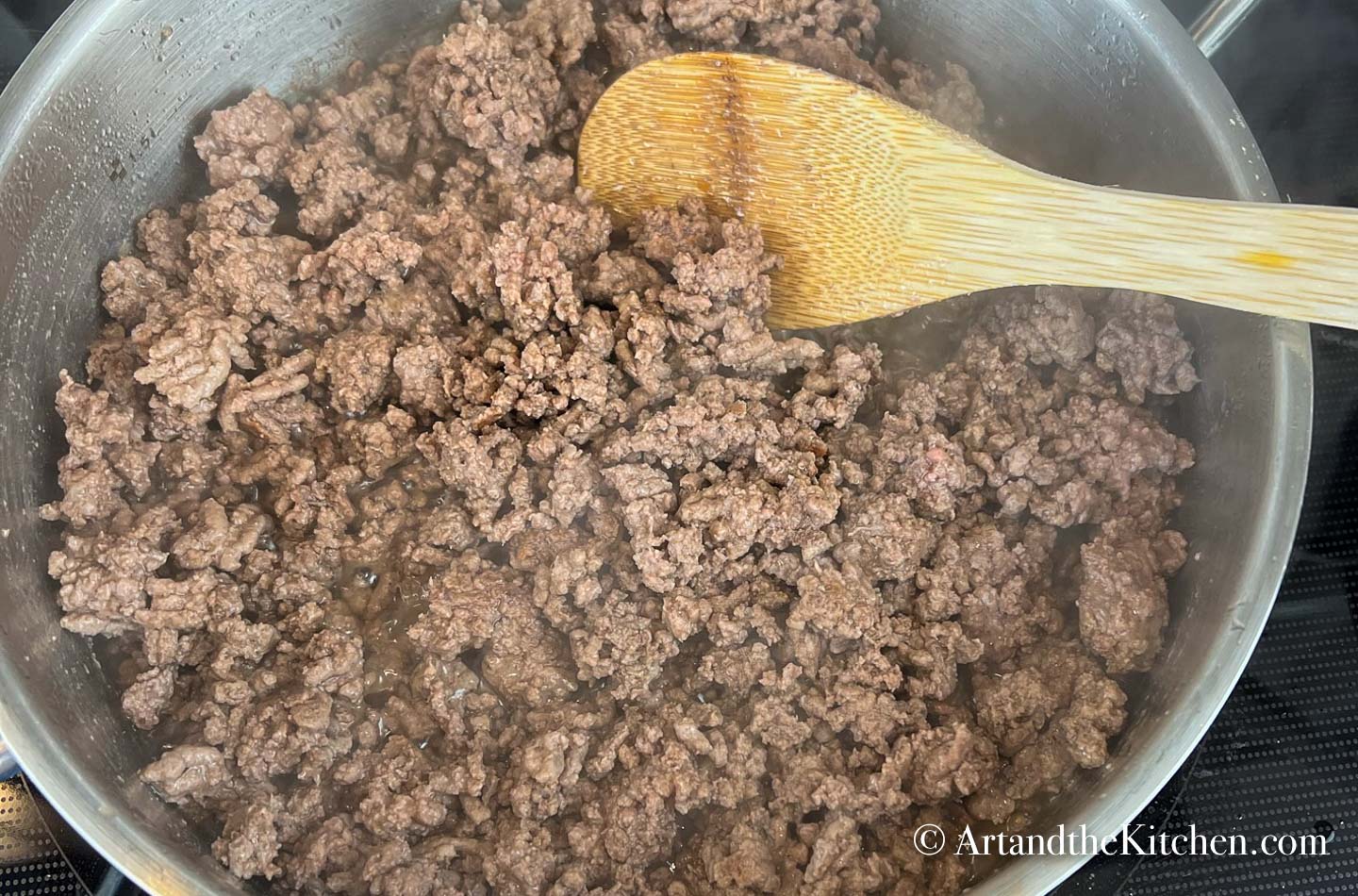 Stainless steel skillet filled with cooked ground beef. Wooden spoon stirring.
