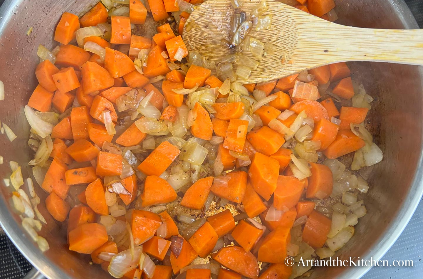 Carrots, onions and garlic cooking in stainless steel skillet. Wooden spoon stirring mixture.