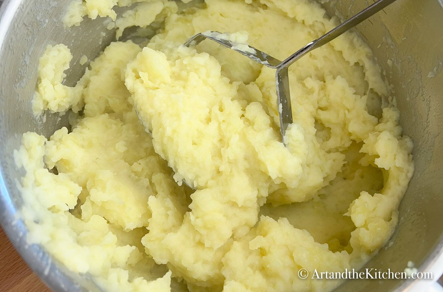 Potato masher mashing potatoes in stainless steel pot.