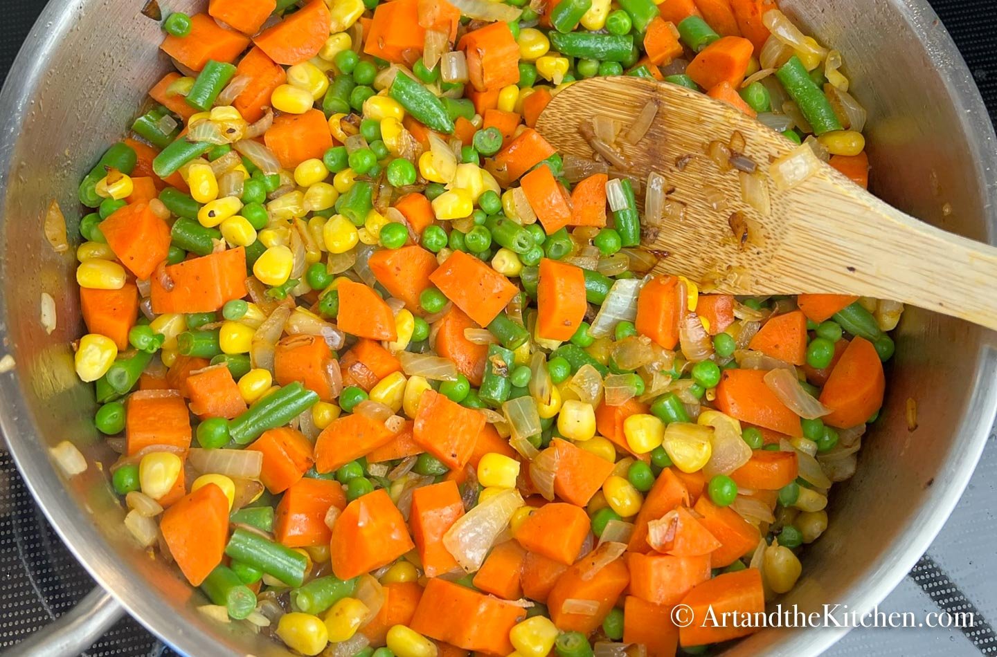 Stainless steel skillet filled with cooked carrots, green beans, peas and onions. Wooden spoon stirring mixture.