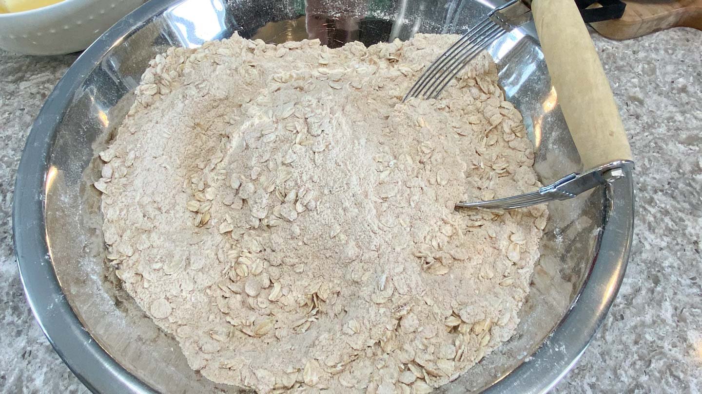 Stainless steel bowl of flour, oats, brown sugar, pastry blender on side.