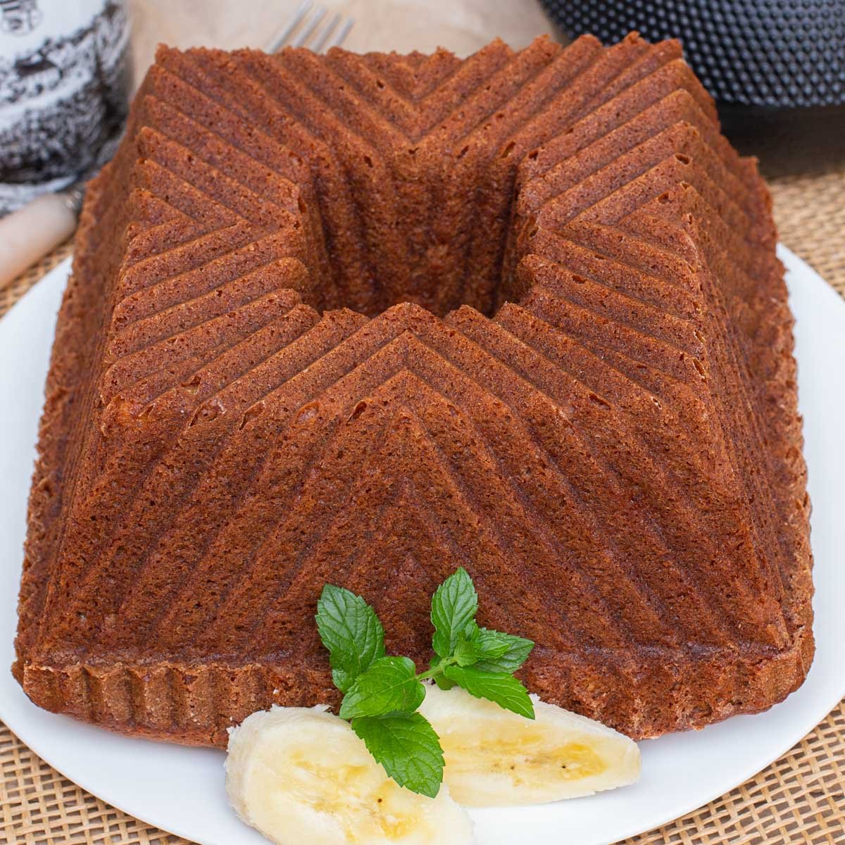 Banana bread on white plate, made in decorative square bundt cake pan, garnished with mint and sliced bananas.