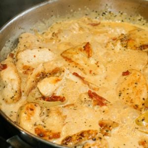 Chicken with shallots and sun-dried tomatoes in a creamy sauce simmering in a stainless steel frying pan.