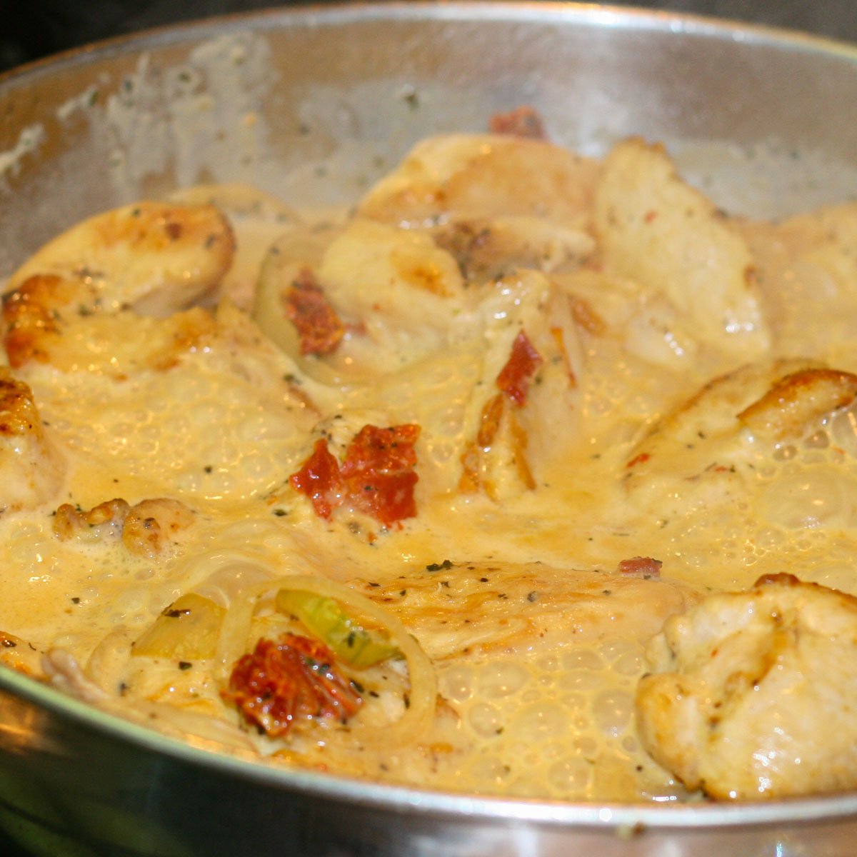 Chicken with shallots and sun-dried tomatoes in a creamy sauce simmering in a stainless steel frying pan.