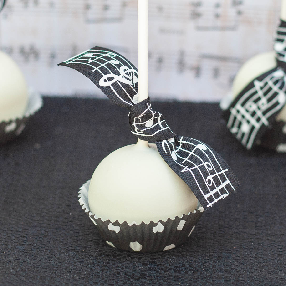 White chocolate coated cake pop in black paper baking cup, tied with music themed ribbon.