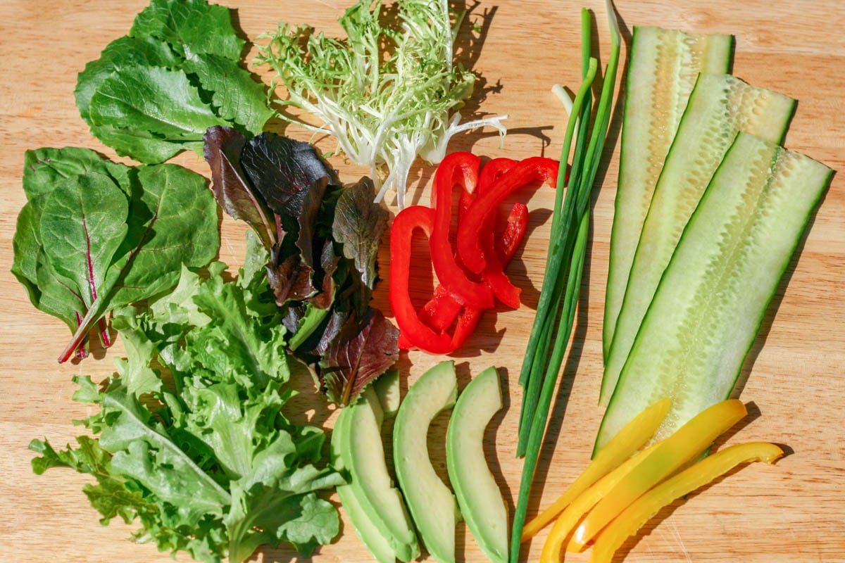 Salad greens, cucumber, red and yellow pepper slices and avocado slices on wood cutting board.