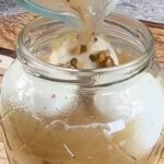 Brine being poured over hard boiled eggs in glass jar and measured ingredients displayed.