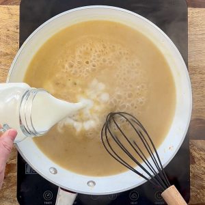 Milk pouring into brown sauce in white skillet with black whisk.