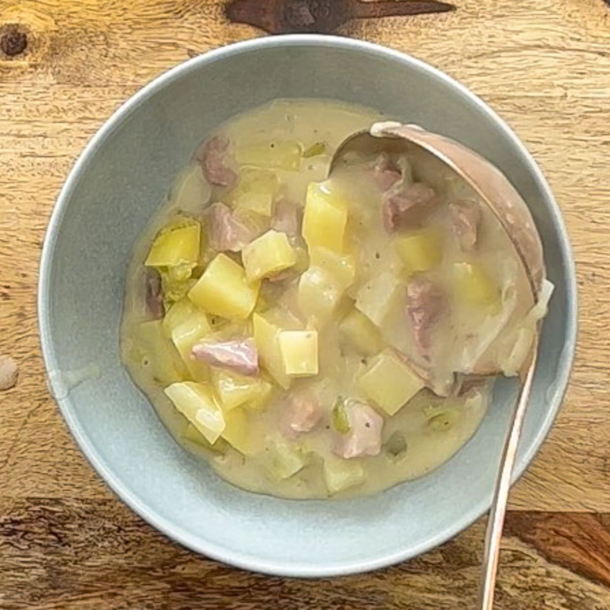 Ladle pouring potato soup with ham into a blue bowl.