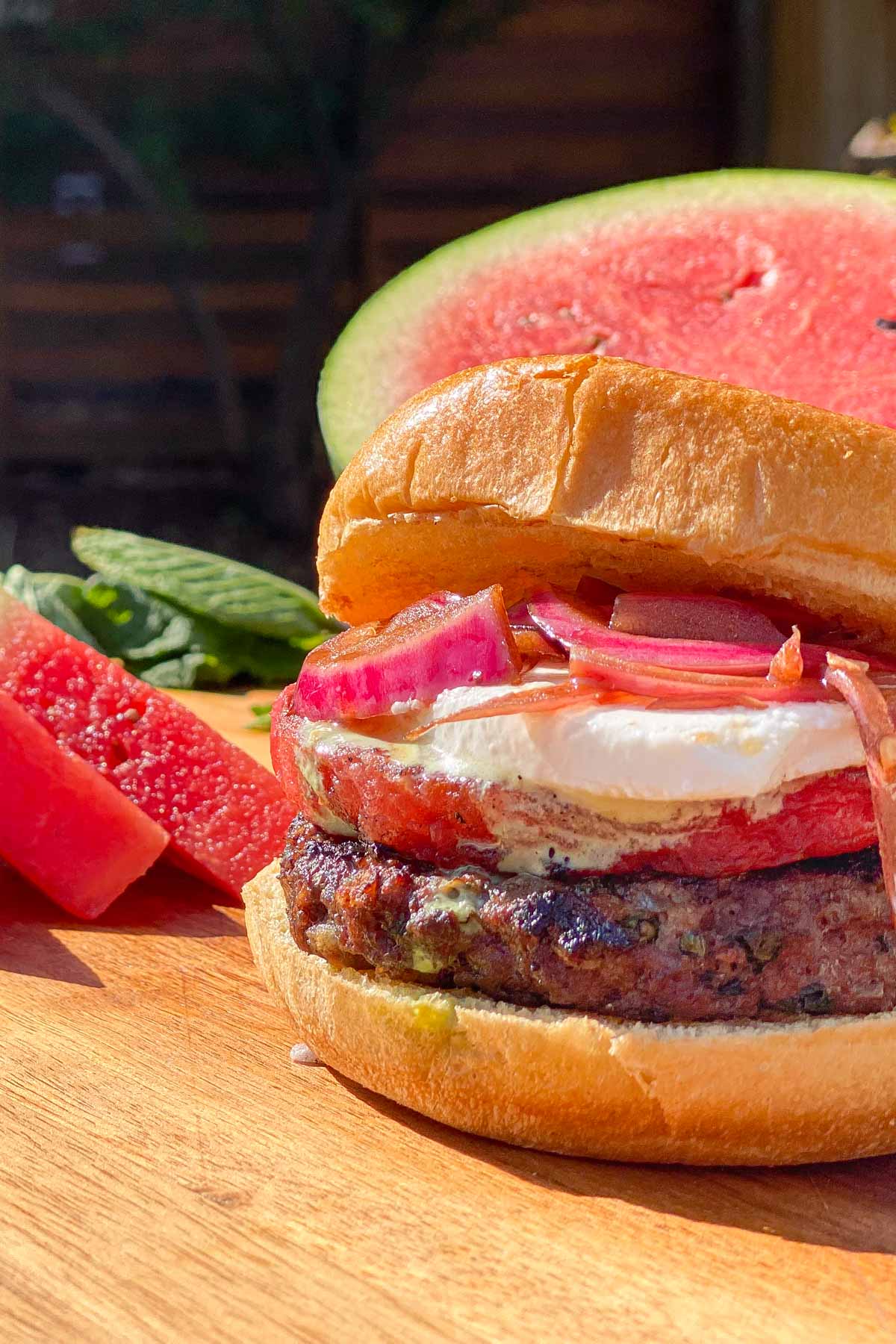 Grilled hamburger with watermelon, feta cheese, and red onions, sliced watermelon in background.