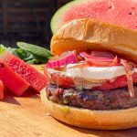 Grilled hamburger with watermelon, feta cheese, and red onions, sliced watermelon in background.