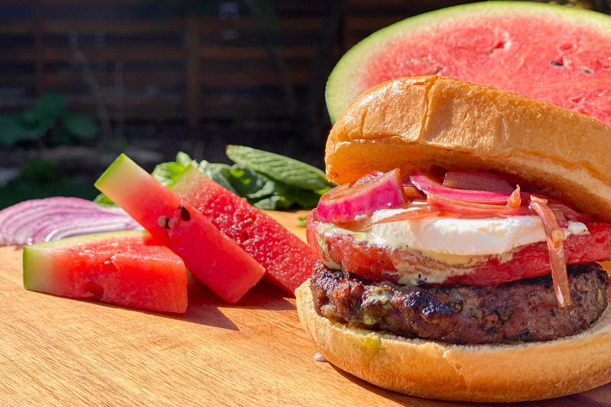Grilled hamburger with watermelon, feta cheese, and red onions, sliced watermelon in background.