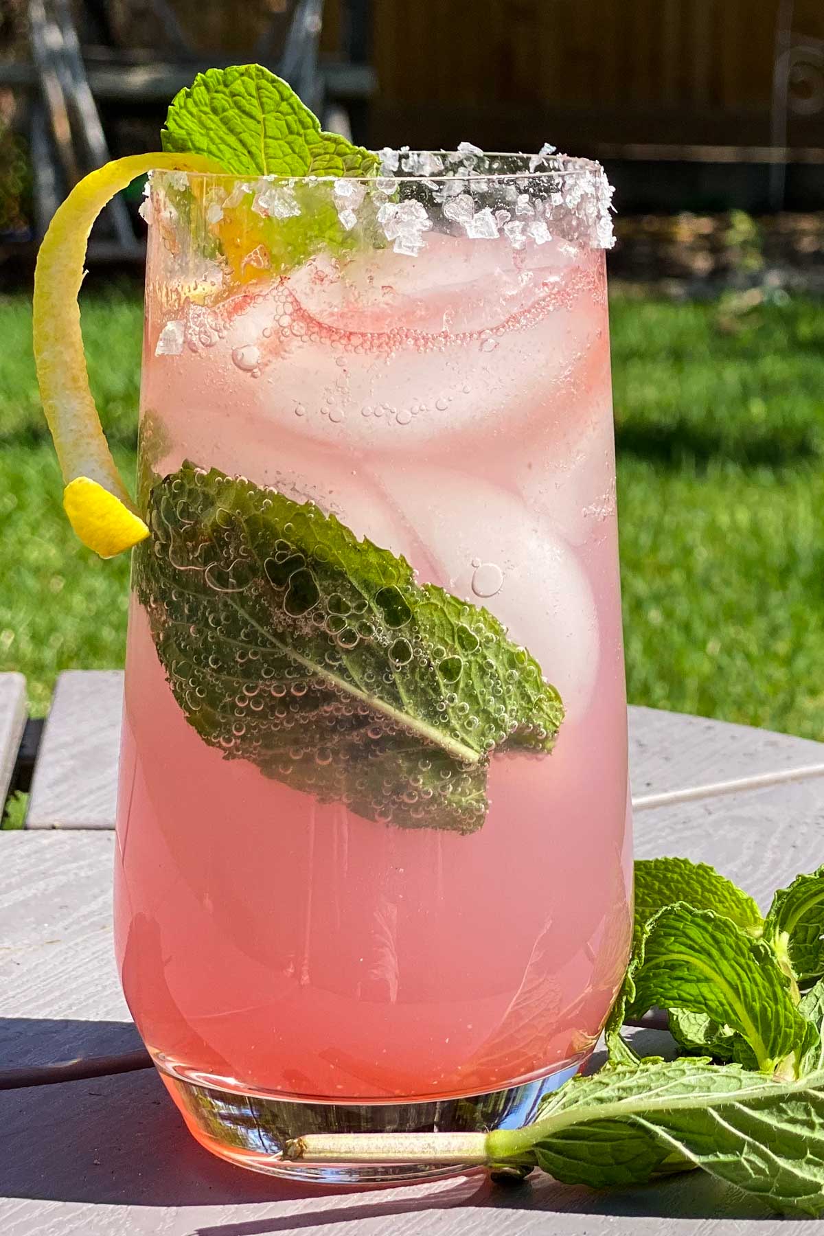 Tall glass filled with pink cocktail, garnished with lemon curl and mint leaves.