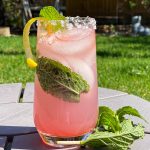 Tall glass filled with pink cocktail, garnished with lemon curl and mint leaves.