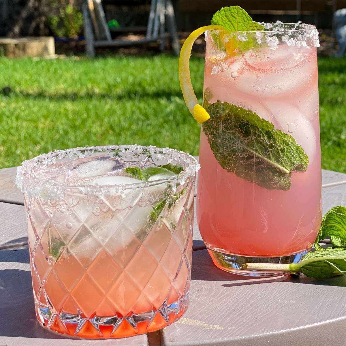 Tall and short glasses filled with gin cocktail, garnished with lemon curl and mint leaves.