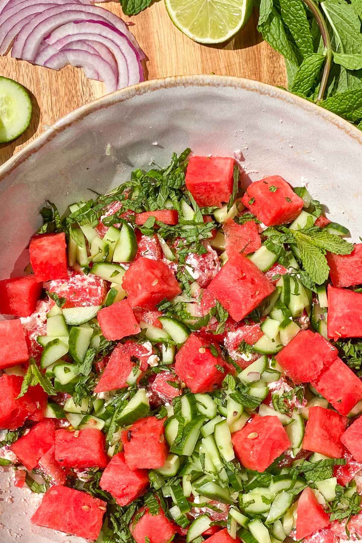 Bowl of watermelon, cucumber, mint, and feta cheese salad with a mint sprig, lime and slices of red onion in background.