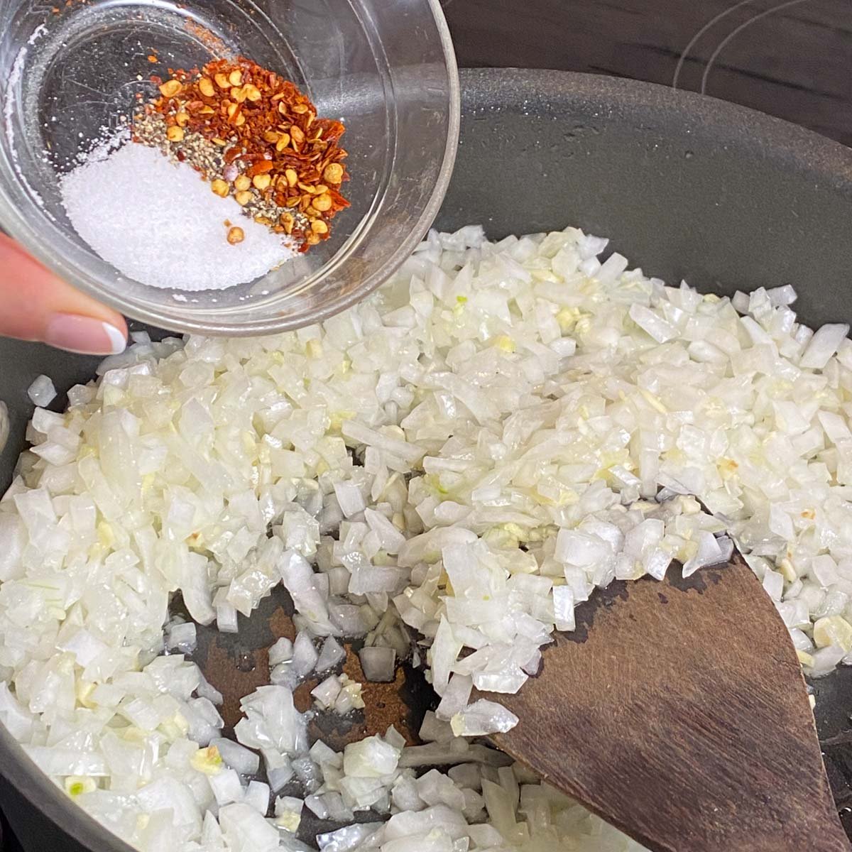 Skillet of sautéed onion, with small bowl of red pepper flakes, salt and pepper being pouring on top.