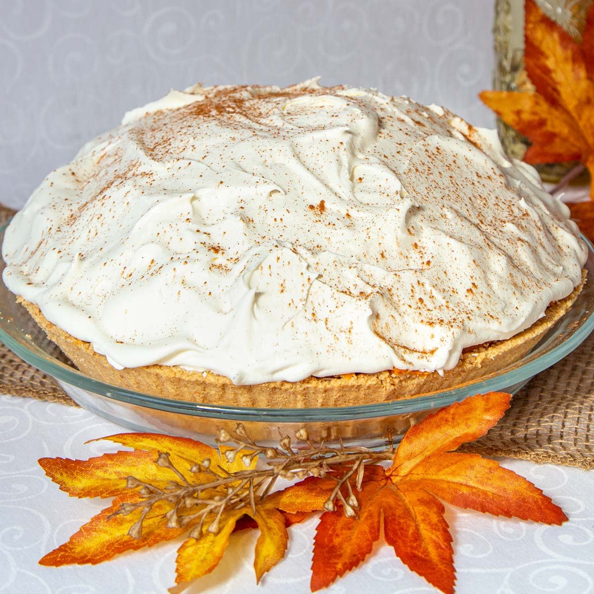 No bake triple layer pumpkin pie with graham crust topped with whipping cream. Decorative fall leaves in background.